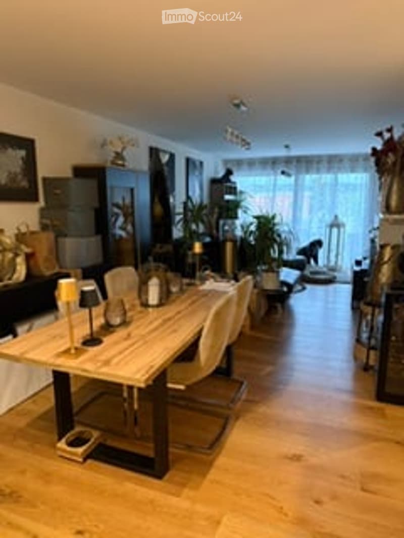 Bright, modern dining area with a large wooden dining table surrounded by chairs, candle holders, and decorative vases. The space features wooden flooring, large windows with sheer curtains, and additional seating in the background.