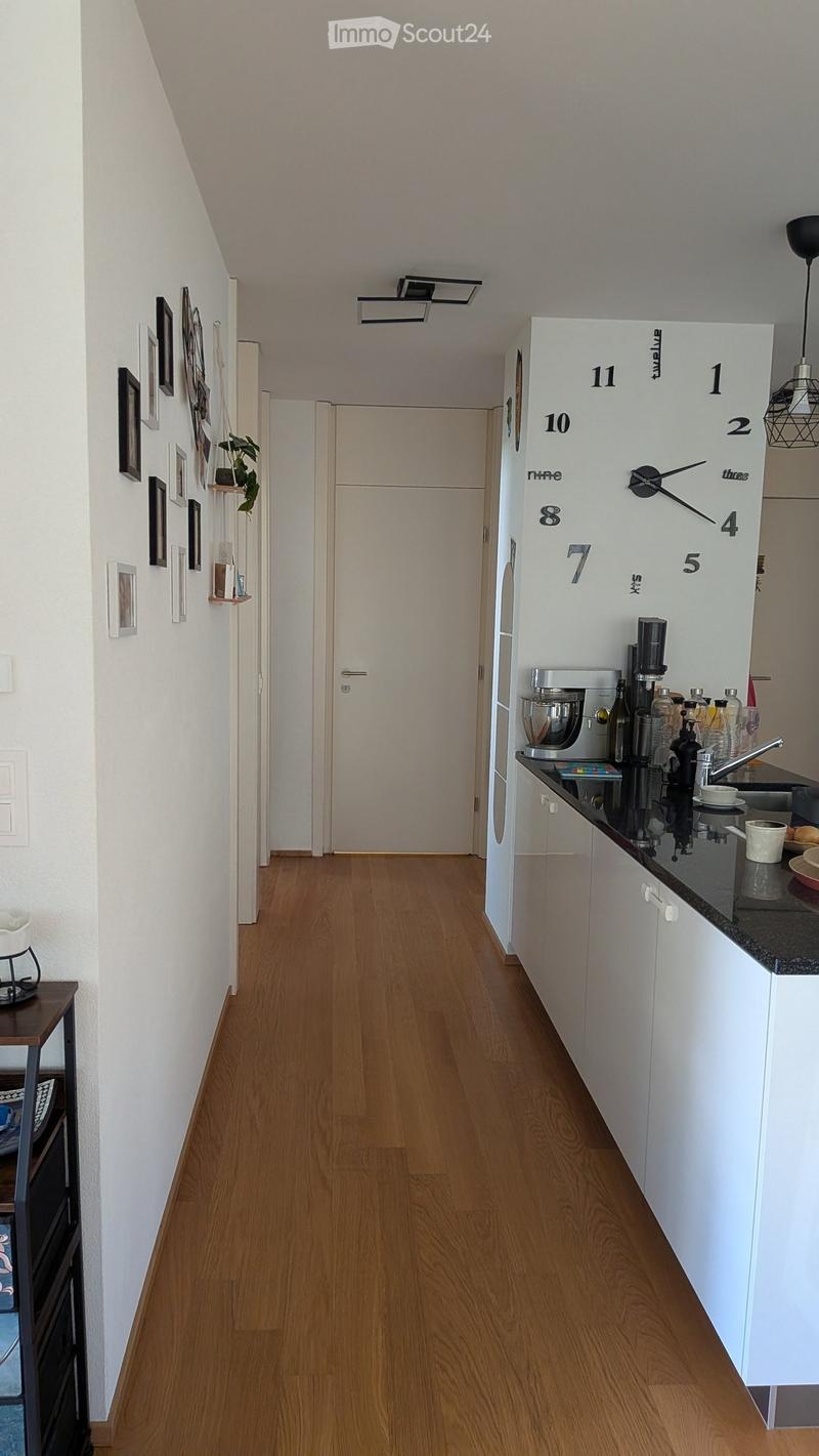 A narrow hallway with wooden flooring, white walls, and a small kitchen area visible on the right side. The kitchen features white cabinets, a countertop with various appliances, and a clock on the wall. A door is visible at the end of the hallway.
