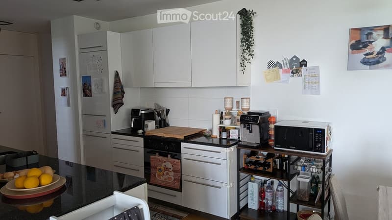 Modern kitchen with white cabinets, stainless steel appliances, and a black countertop. Includes a refrigerator, oven, microwave, coffee maker, and a wine rack with bottles. A bowl of oranges and a cutting board are on the counter.