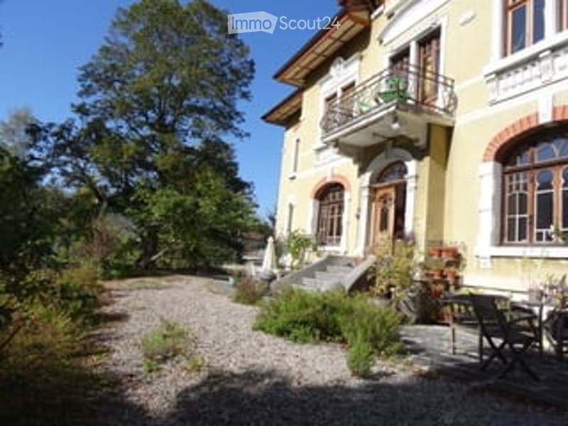 A large, yellow villa with arched windows and a balcony, surrounded by greenery and a gravel driveway.