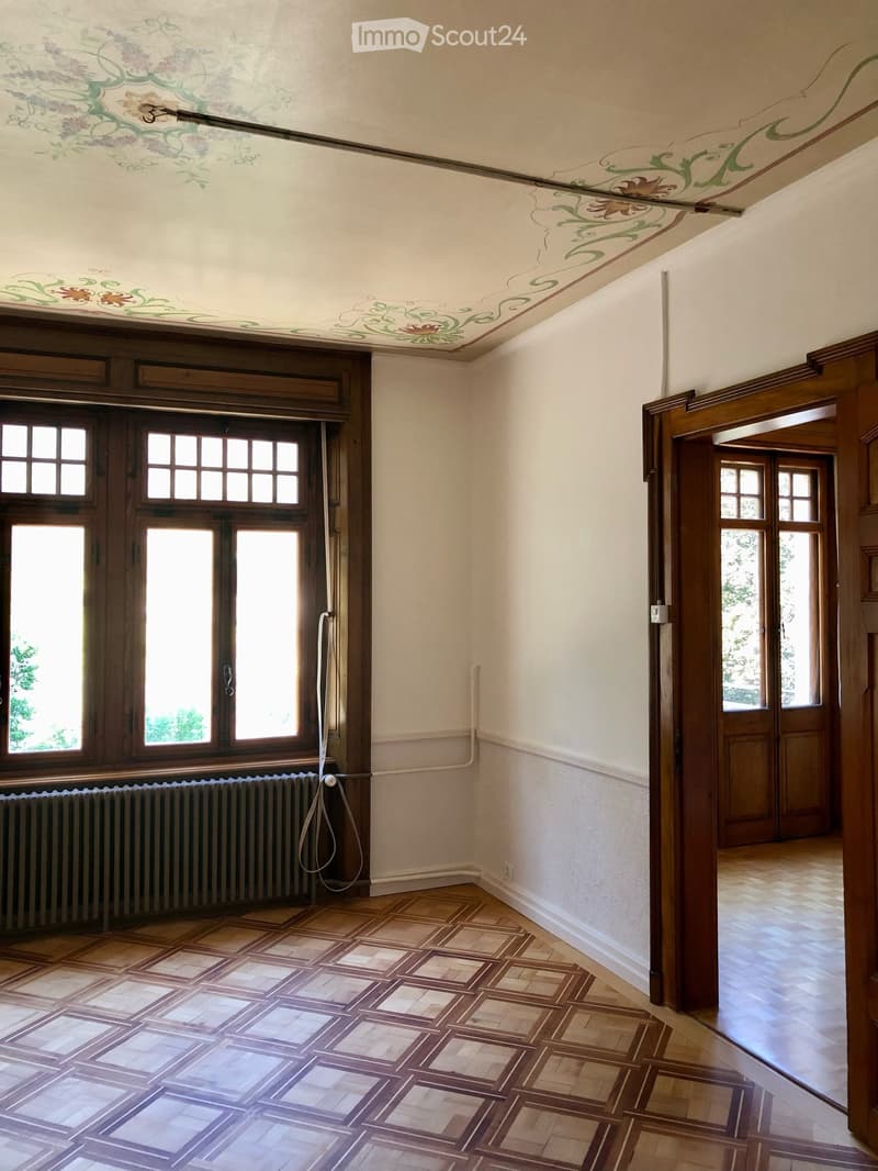 An empty room with a decorative painted ceiling, wooden doors, large windows, and a patterned tile floor.