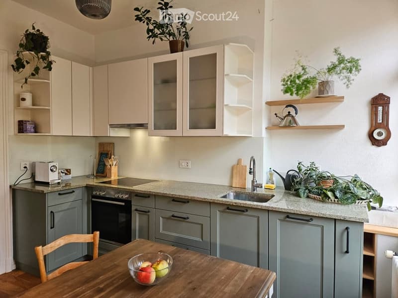 Modern kitchen with light-colored cabinets, grey lower units, a wooden dining table with a bowl of fruit, and various appliances including a toaster, oven, and sink.