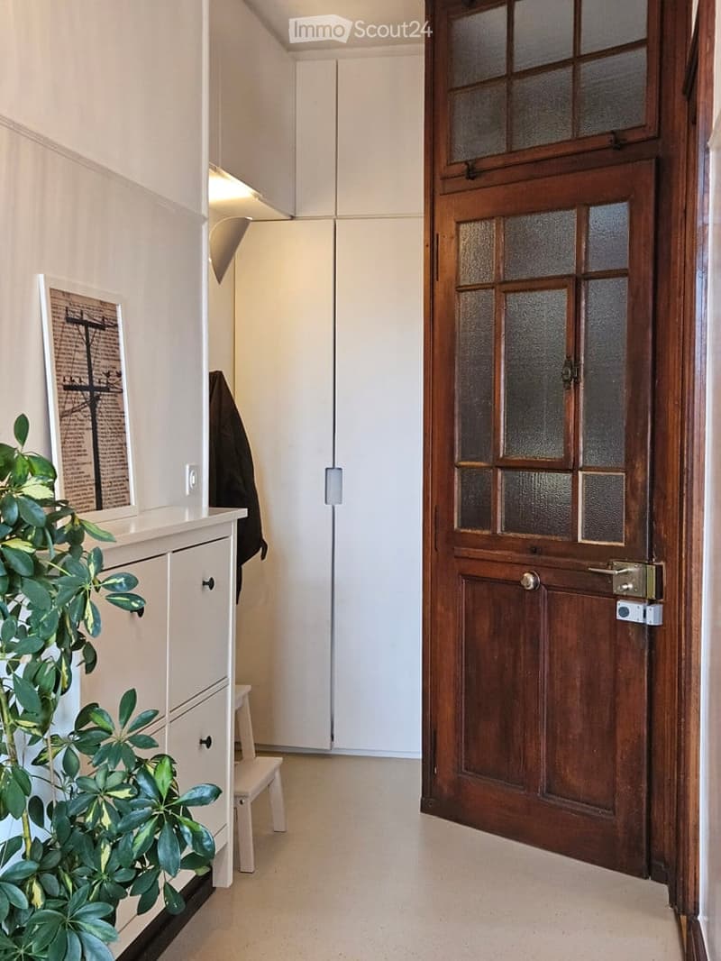 A modern hallway with a wooden door featuring frosted glass panels, a white wardrobe, a small plant, and a framed picture on a console table.