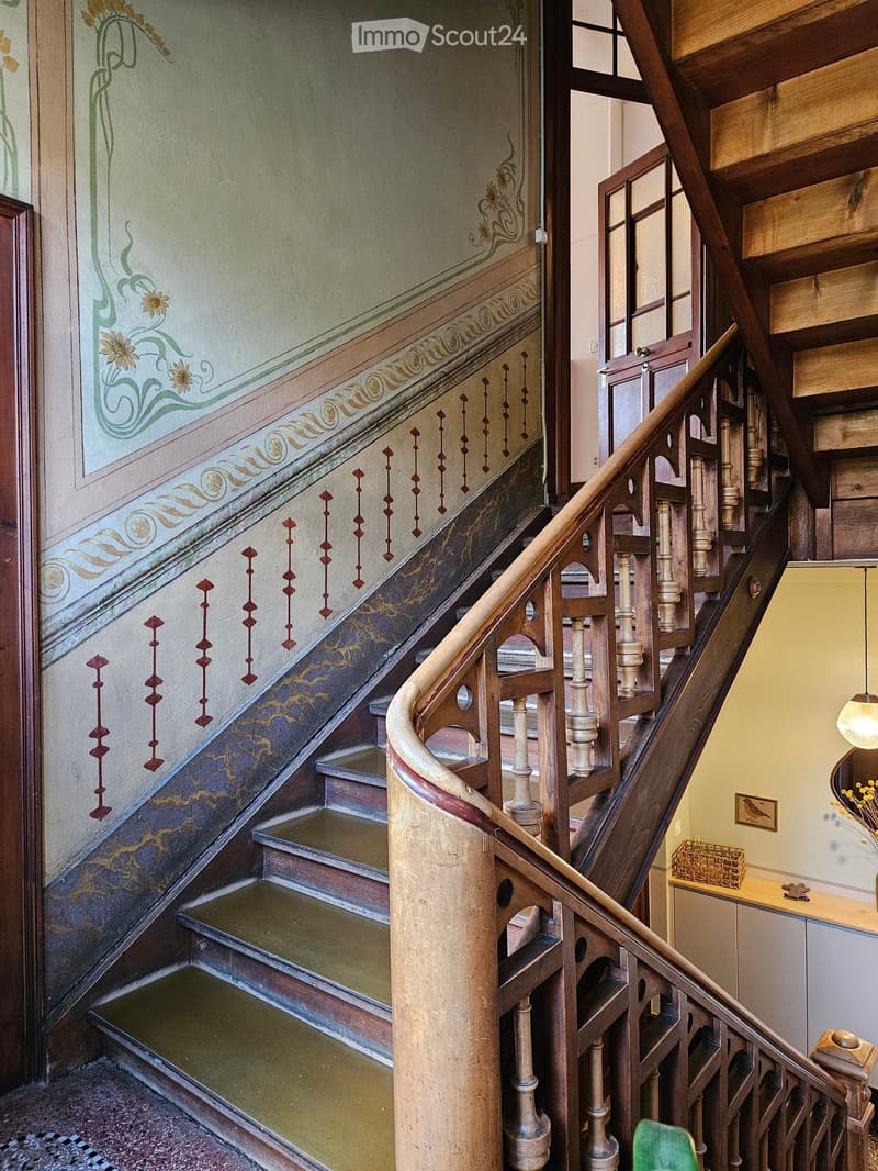Ornate wooden staircase with decorative railings and painted walls featuring floral and geometric patterns. The steps are carpeted, and the walls have intricate designs and artwork.