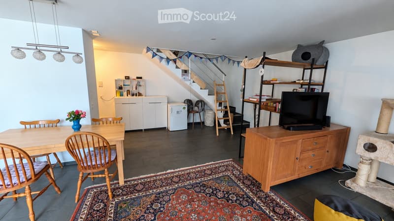 A well-lit, multi-functional room with a dining area, a TV area, and a small kitchen. Features include a wooden dining table with chairs, a TV on a wooden cabinet, a cat tree, a small refrigerator, and a staircase leading to another level.