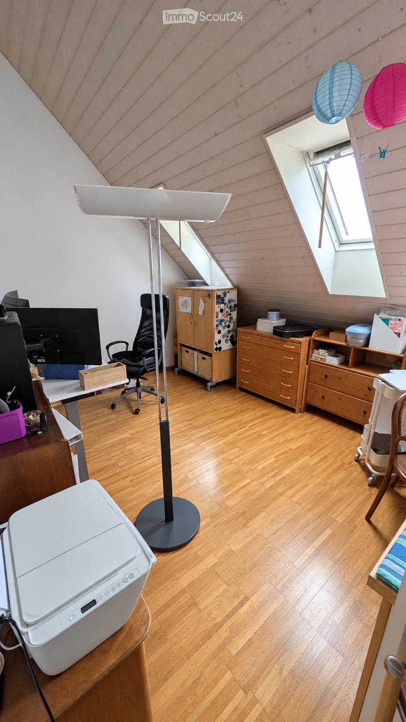 A well-lit attic room with wooden flooring, featuring a desk with a chair, a large screen, a printer, a tall floor lamp, and wooden storage units including cabinets and shelves.