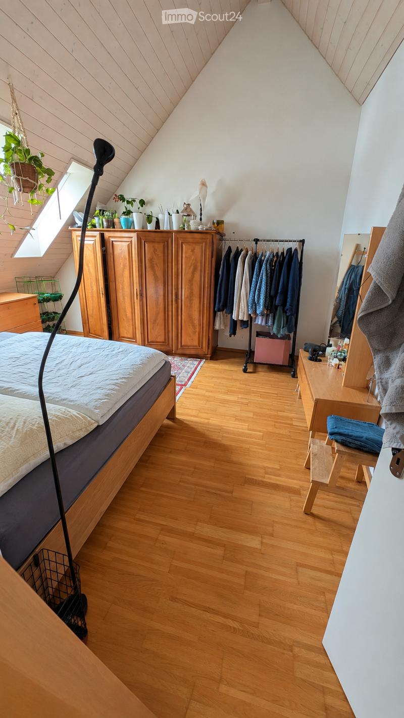 Cozy attic bedroom with wooden floors, slanted wooden ceiling, large window, bed, wardrobe, mirror, small table, and hanging plants.