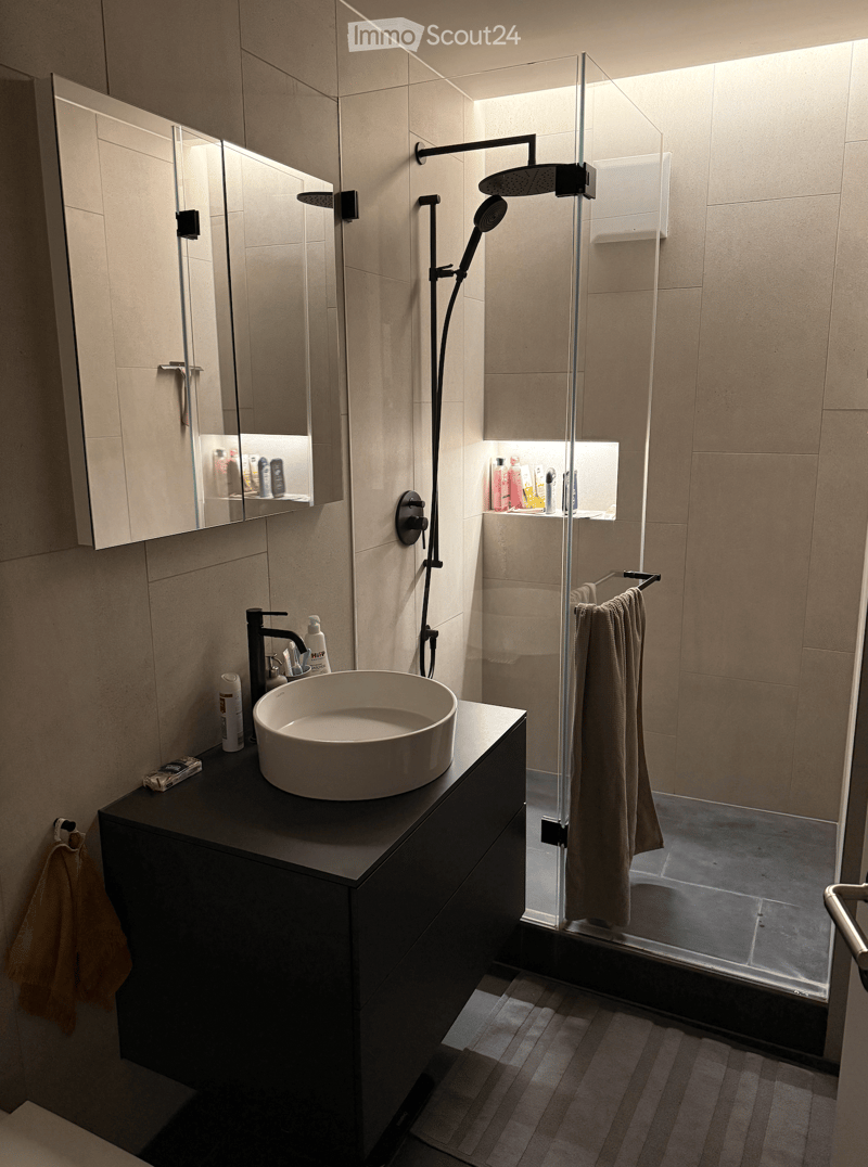 Modern bathroom with a sleek black vanity, white sink, large mirror, and a glass-enclosed shower with a modern showerhead. Beige tiles and warm lighting create a contemporary ambiance.