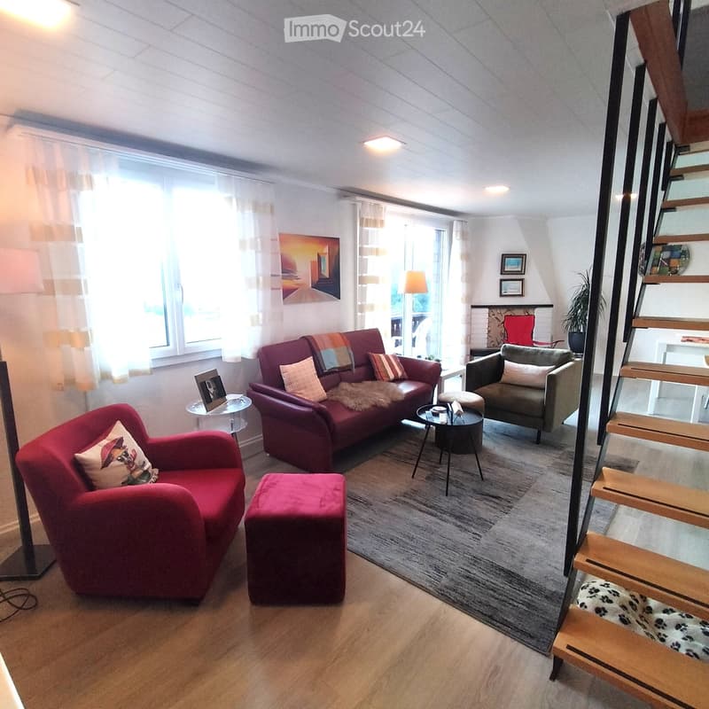 Modern living room with a red sofa, grey armchair, ottoman, coffee table, side table, and decorative pillows. Large windows with sheer curtains and a staircase leading to an upper level.