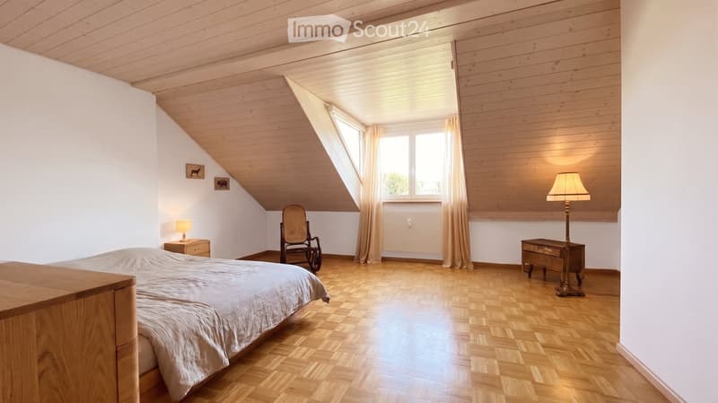 Cozy attic bedroom with wooden floors, slanted wooden ceiling, large windows with curtains, a bed, nightstand, lamp, rocking chair, and a small table with a lamp.