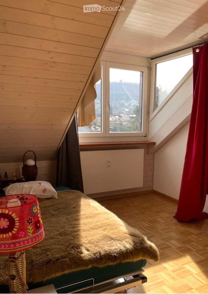Cozy attic bedroom with wooden slanted ceilings, large windows offering a scenic view, a neatly made bed with a furry throw, a colorful lamp, and a wicker basket on a nightstand.