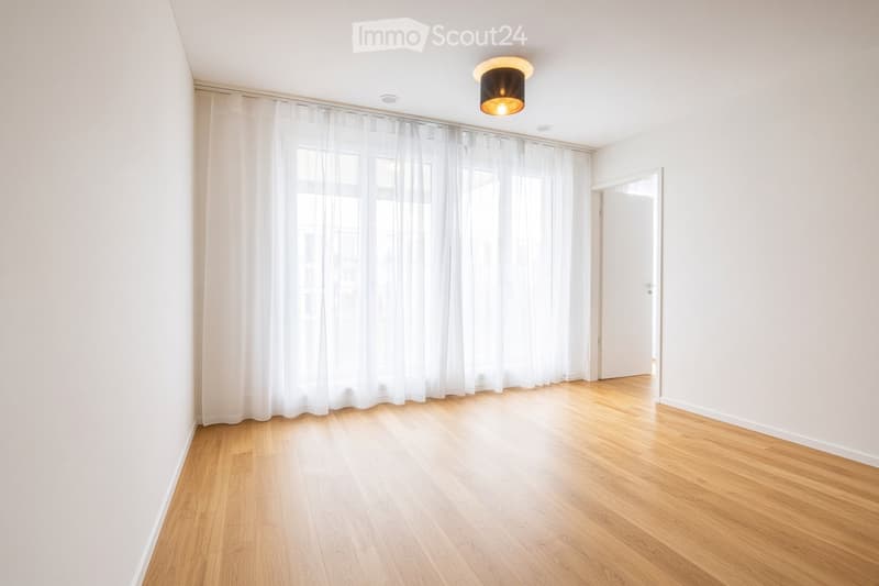 Empty room with wooden flooring, white walls, large window with white curtains, and a single ceiling light fixture.