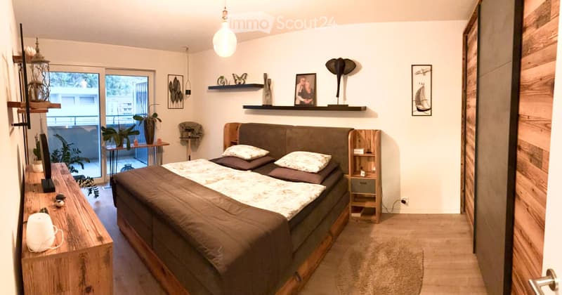 Modern bedroom with a large bed, nightstand, wooden table, chair, shelves with decorative items, and a view of a balcony.
