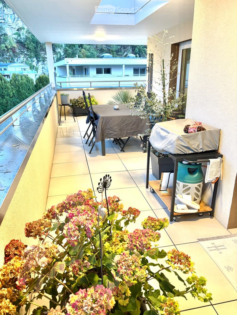 Balcony with tiled floor, table and chairs, flower pots, barbecue, and a view of the surrounding area.