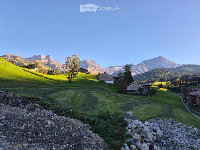 A scenic mountain valley with green hills, scattered wooden houses, and a clear blue sky. The foreground shows a rocky terrain, while the background features snow-capped mountains and a lone tree.