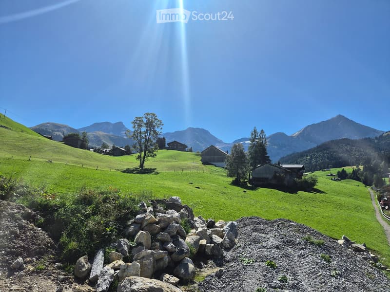 A picturesque alpine landscape with green hills, scattered houses, and distant mountains under a clear blue sky.