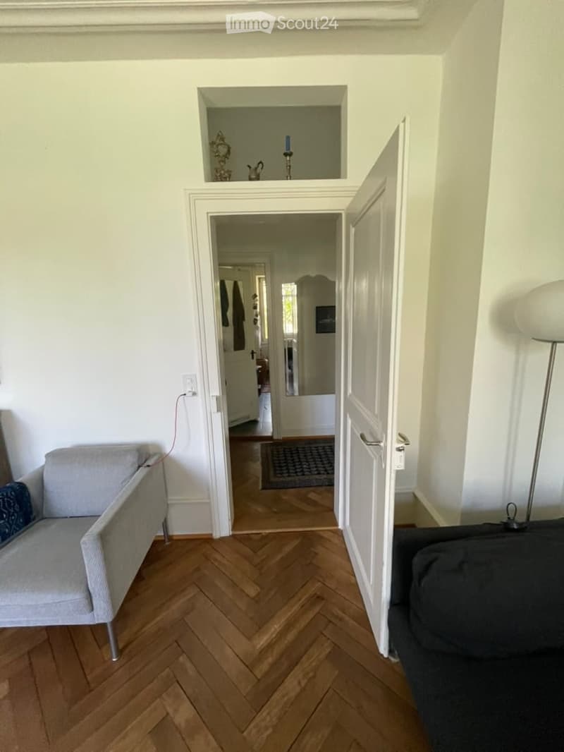 A modern living room with herringbone wooden flooring, white walls, and an open doorway leading to another room. Visible furniture includes a grey sofa, a chair, and a floor lamp.