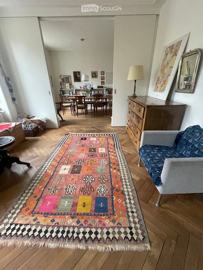 Living room with wooden flooring, colorful rug, sofa, chair, table, lamp, mirror, bookshelf, and picture frames.