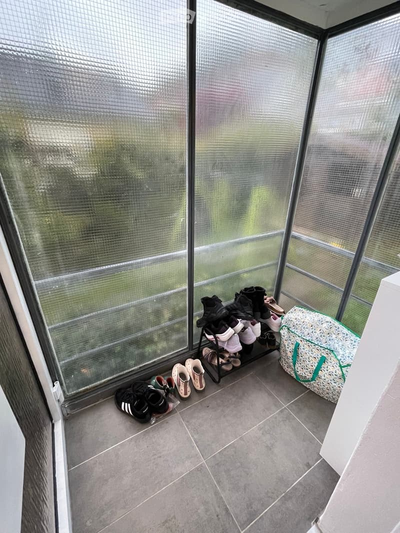 A small, well-lit hallway with large glass windows, a shoe rack holding multiple pairs of shoes, and a green and white patterned bag on the floor.