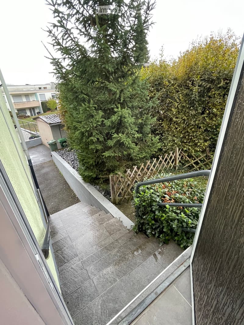 Balcony with tiled floor, metal railing, small wooden fence, and surrounding greenery including trees and bushes.