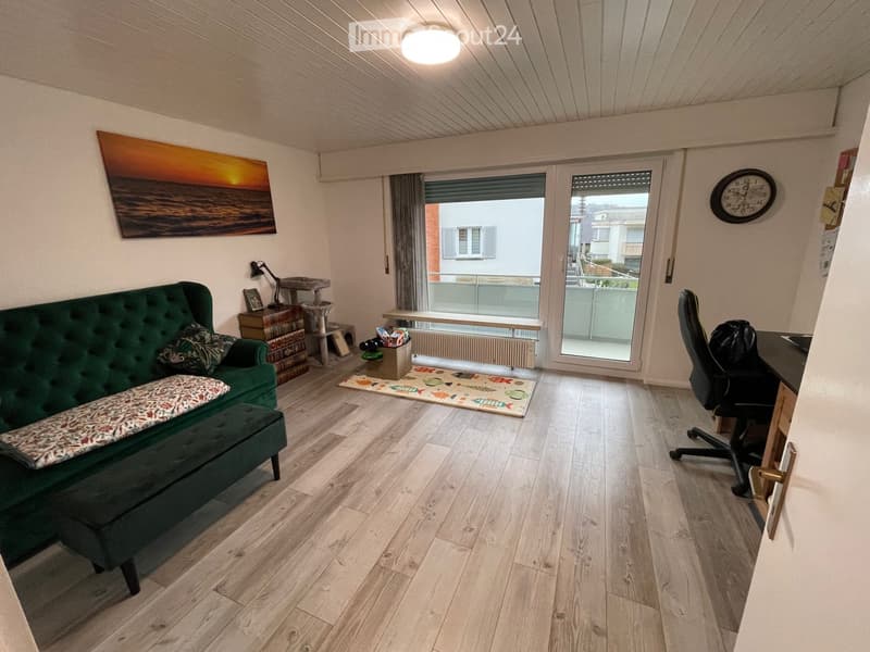 A modern living room with a green sofa, ottoman, wooden floor, a cat tree, a desk with a chair, and a balcony view.