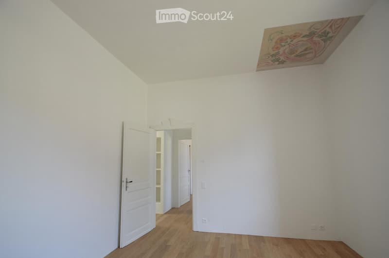 Empty room with white walls, wooden flooring, and a decorative ceiling piece. There is a doorway leading to another room.