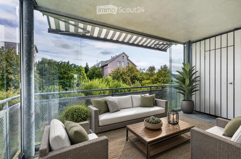 Modern balcony with large windows, offering a scenic view of trees and houses. Features a white wicker sofa, wooden coffee table, two wicker chairs, a potted plant, and a candle holder.