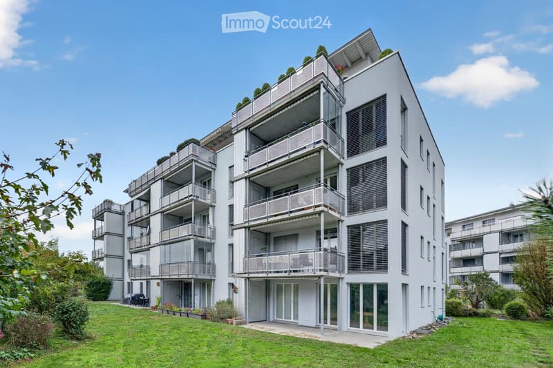 Modern multi-story residential building with white facade, multiple balconies, and surrounding greenery.