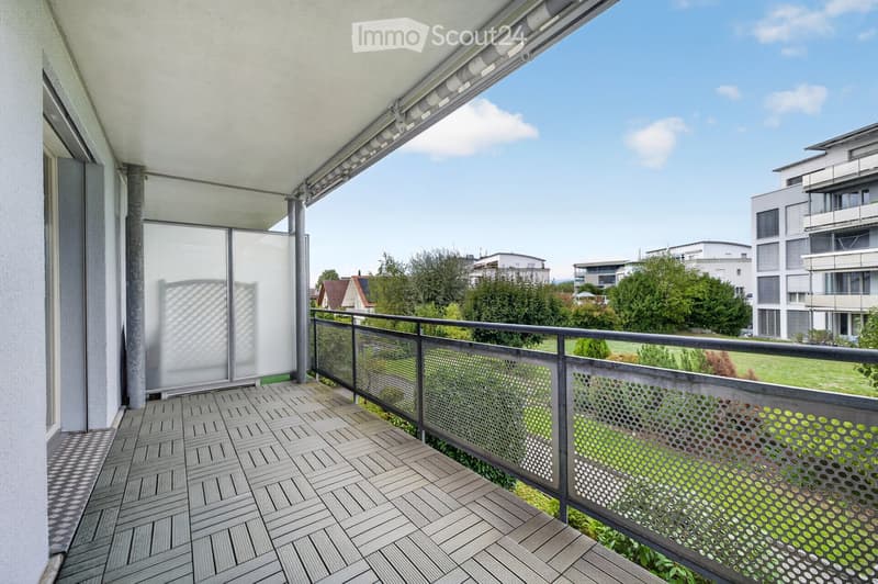 Balcony with wooden decking, metal railing, and a view of a green landscape and modern buildings.