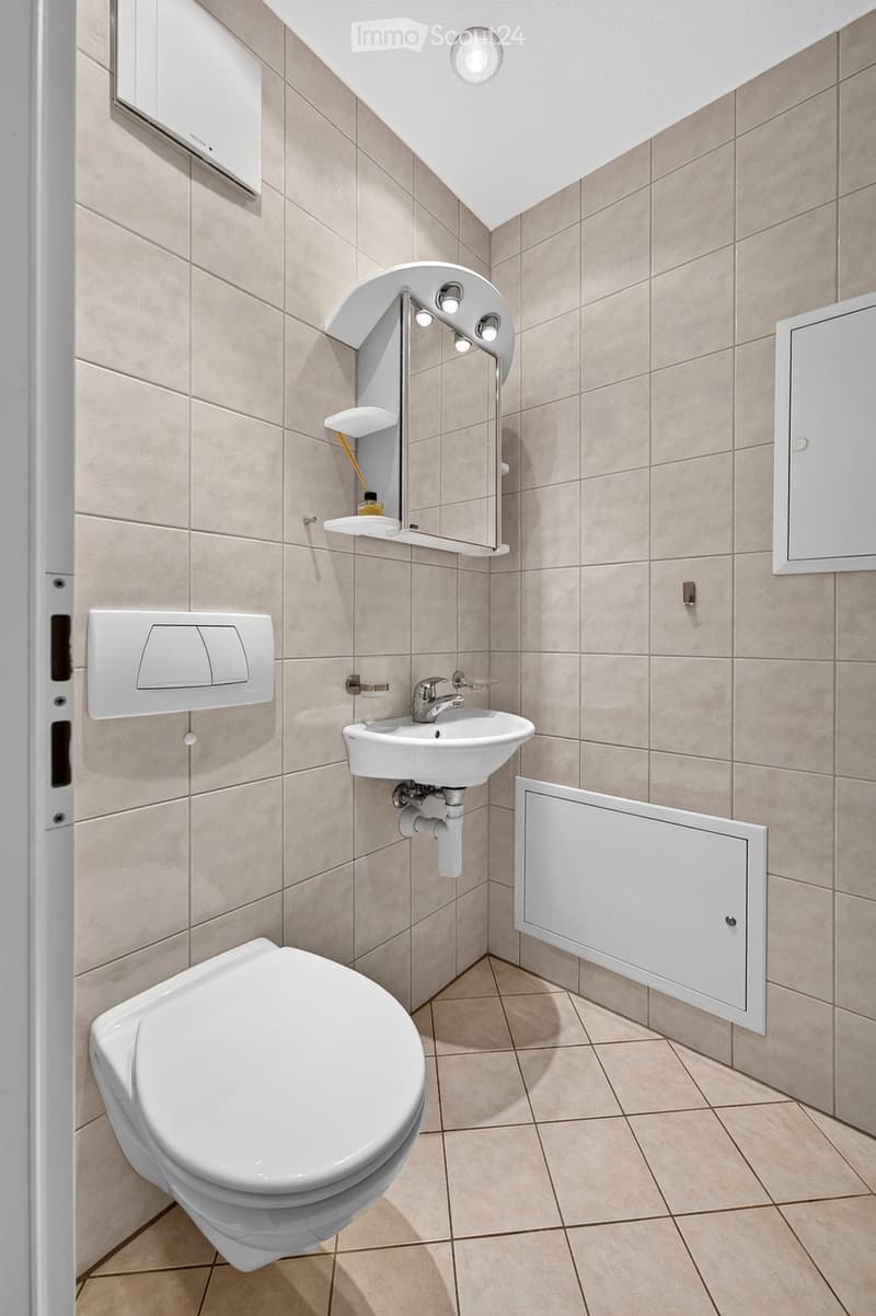 Modern bathroom with beige tiled walls and floor, featuring a toilet, sink with a modern faucet, a mirror with built-in lighting, and a cabinet.