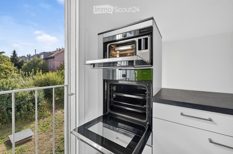 Modern kitchen with built-in ovens, white cabinets, and a view of the garden from the balcony.