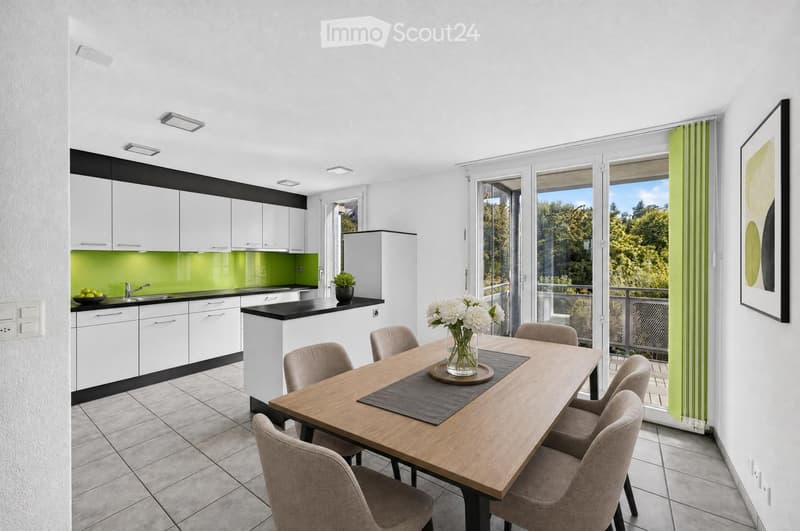 A modern kitchen and dining area with white cabinets, a green backsplash, a wooden dining table with six chairs, and a vase of white flowers on the table. The kitchen features a sink, faucet, and a small potted plant on the counter.