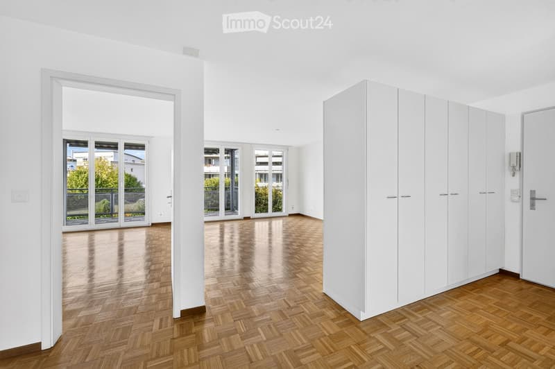 Empty room with white walls, wooden herringbone flooring, large sliding glass doors leading to a balcony, and built-in white closet units.