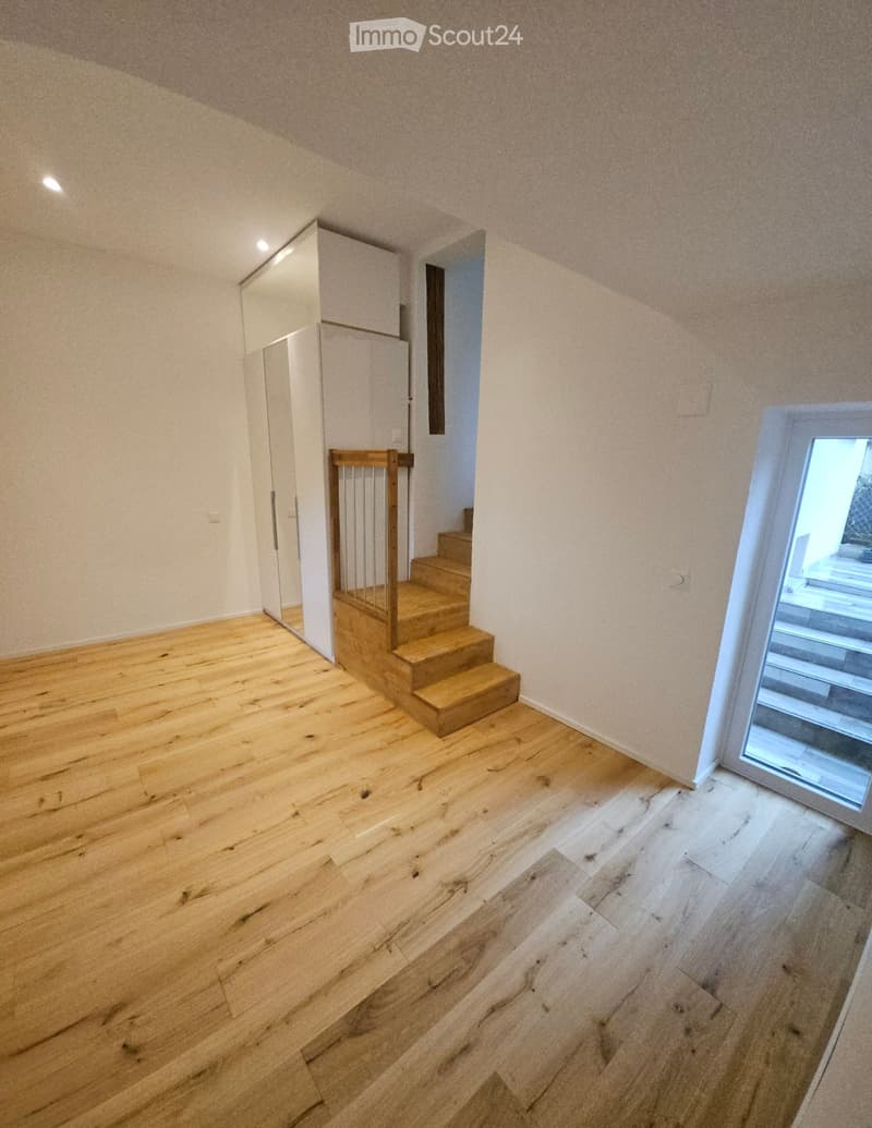 Empty hallway with wooden flooring, white walls, and a staircase leading upwards. A small window on the right side allows natural light to enter.