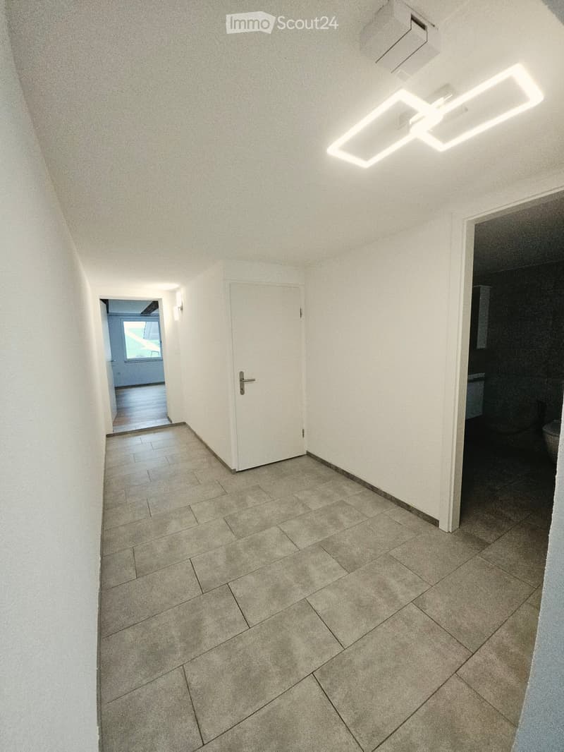Empty hallway with tiled floor, white walls, and a ceiling light fixture. Doors lead to other rooms, including a bathroom and a room with a window.