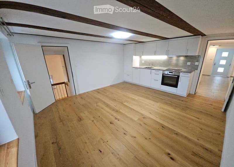 Empty room with wooden flooring, white walls, and exposed wooden beams on the ceiling. A small kitchen area is visible in the background with white cabinets and appliances.