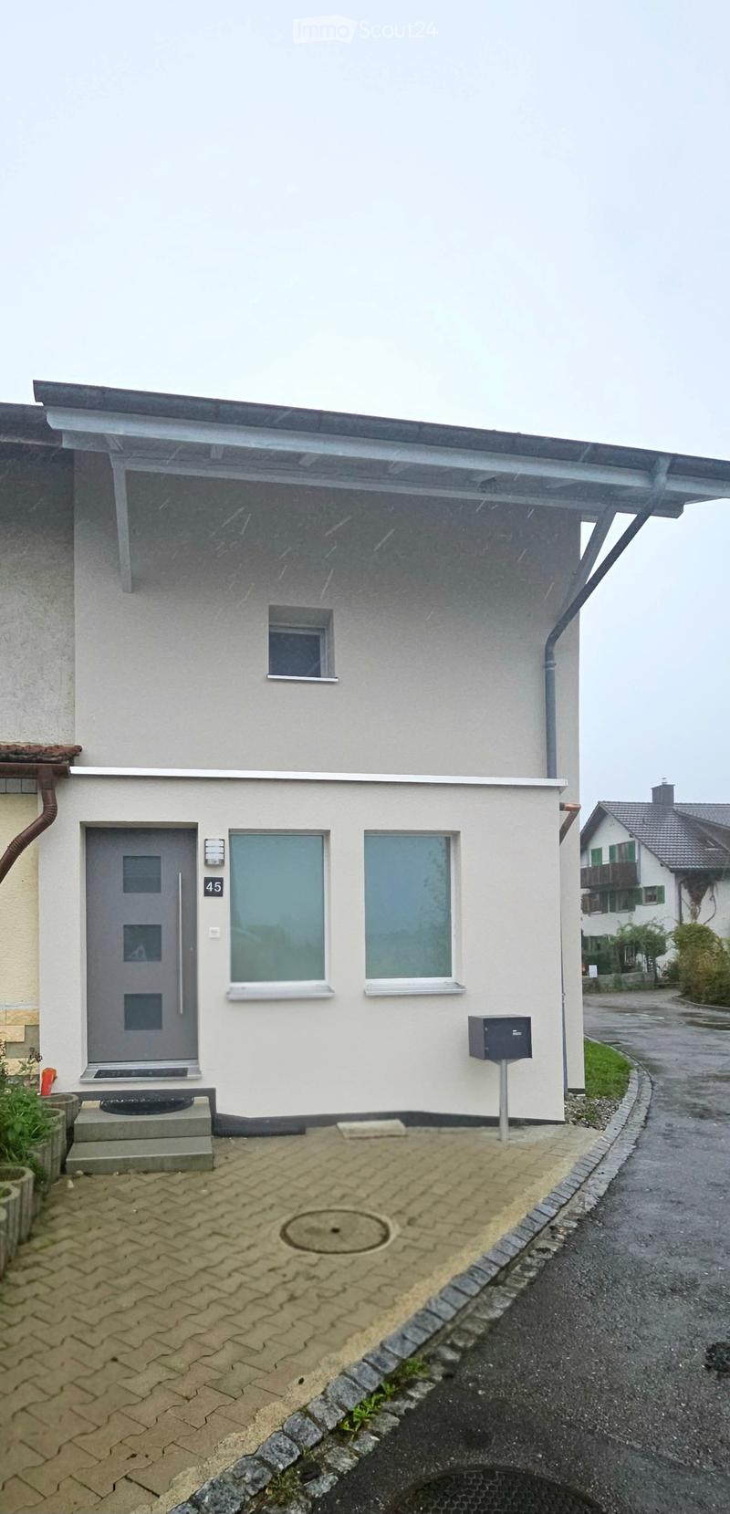 Single-story house with a gray front door, two windows, and a small porch area with a mailbox. The house has a modern design with a flat roof and a small garden area to the left.