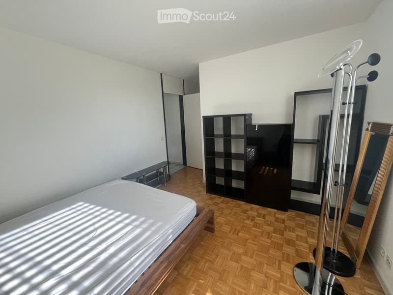 Modern bedroom with wooden flooring, a neatly made bed, a large mirror, a black shelving unit, and a small table.