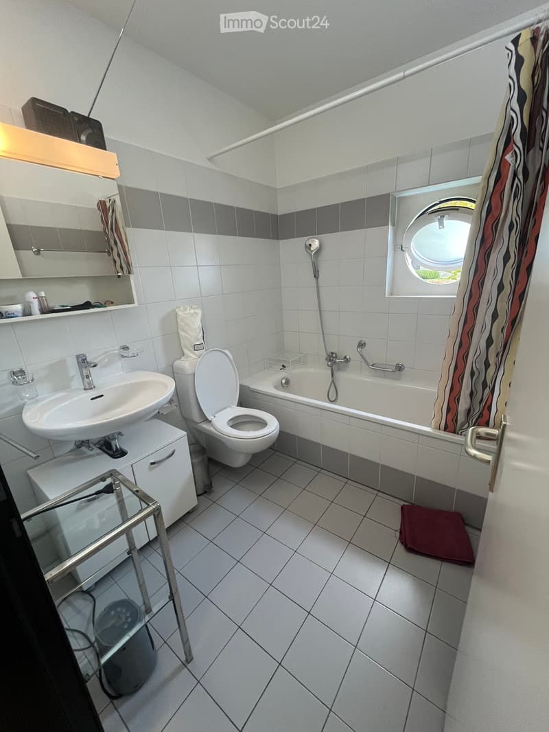 Modern bathroom with white tiles, a sink, toilet, bathtub, mirror, and a round window. Includes a glass table and a red bath mat.