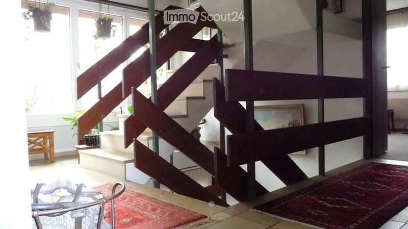 Modern interior with a wooden staircase, a red area rug, a sofa, a small table, and potted plants near the windows.