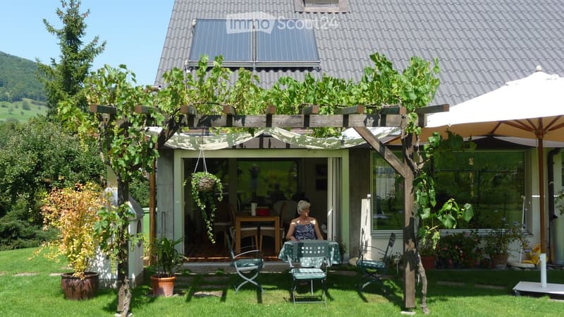 Garden area with a pergola, outdoor seating, and a view of a house with solar panels on the roof. There are potted plants, a hanging basket, and a large umbrella providing shade.