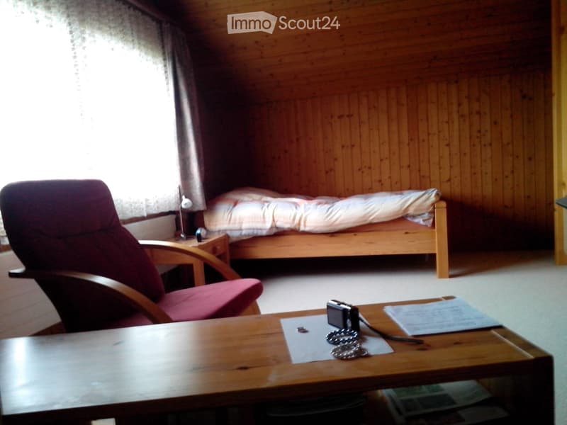 Cozy bedroom with wooden walls, a single bed, a red armchair, a wooden table with a camera and papers, and a small nightstand with a lamp.