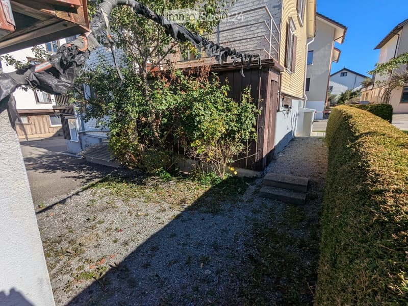 Narrow pathway lined with gravel, small steps leading up, surrounded by greenery and neighboring houses.