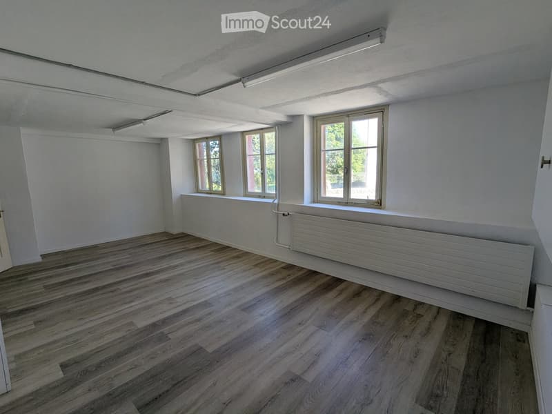 Empty room with wooden flooring, white walls, and large windows letting in natural light. Features a radiator and a door visible on the left side.