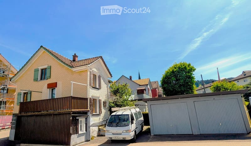 Two-story house with beige and white exterior, green shutters, and a balcony on the upper floor. A van is parked in front of a garage.