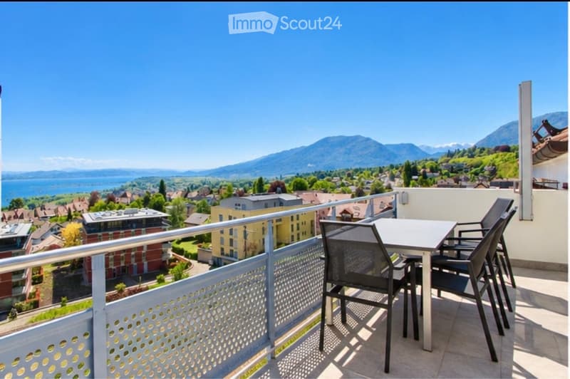 Balcony with a table and chairs, overlooking a scenic view of a town, lake, and mountains.