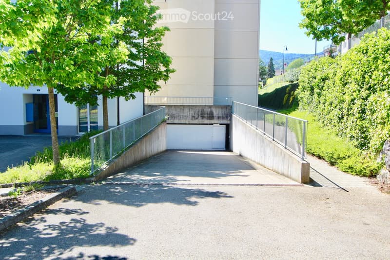 Modern building entrance with a covered driveway leading to a garage, surrounded by greenery and trees.