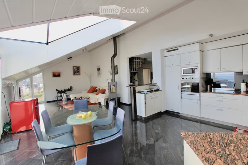 Open-plan living and dining area with a modern kitchen. Features a sofa with orange cushions, a glass dining table with chairs, a red refrigerator, white cabinets, and a fireplace.