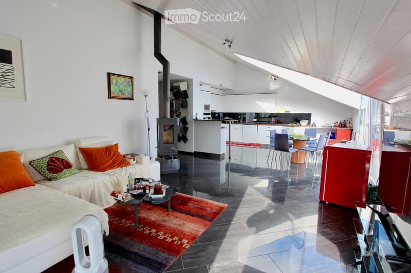 Modern living room with a white sofa, colorful cushions, a coffee table, and a fireplace. The space is open-plan with a kitchen area featuring white cabinets, a red island, and various appliances.