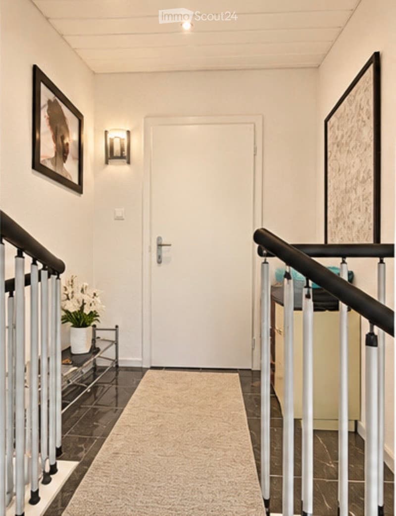 Modern hallway with a white door, black railing, and a small table with a potted plant. The walls are painted white, and there is a light fixture on the wall.